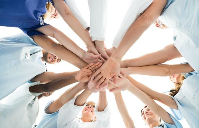 Das Bild zeigt ein motiviertes Team aus Pflegekräften und medizinischem Personal im Kreis, die ihre Hände in der Mitte aufeinanderlegen