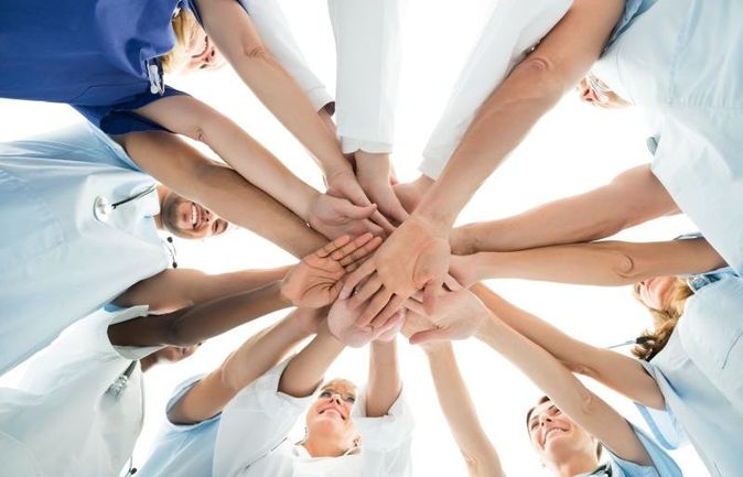 Das Bild zeigt ein motiviertes Team aus Pflegekräften und medizinischem Personal im Kreis, die ihre Hände in der Mitte aufeinanderlegen