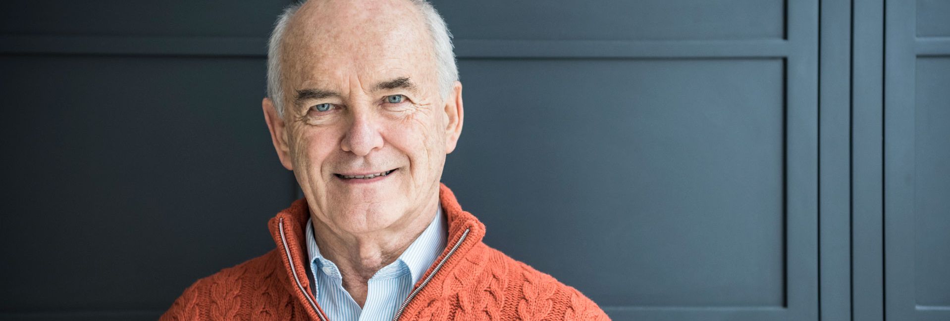 Älterer Mann lächelnd in orange Zopfpullover vor dunkler Wand