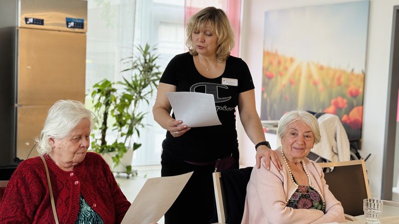 Zwei Gäste sitzen mit Zettel an einem Tisch, dazwischen steht eine Mitarbeiterin und singt mit