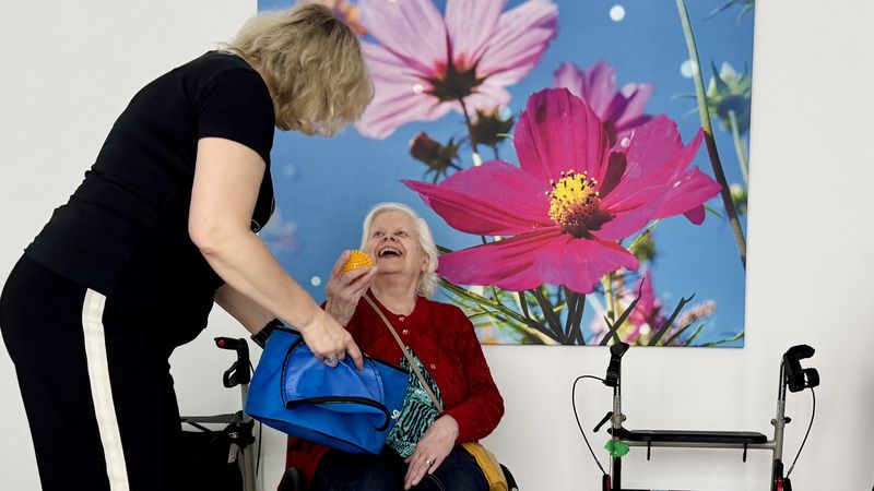 Ältere Frau sitzt auf einem Stuhl. Hinter ihr ist ein großes Leinwandbild mit Blumen zu erkennen. Eine Mitarbeiterin steht neben ihr und spricht mit ihr.