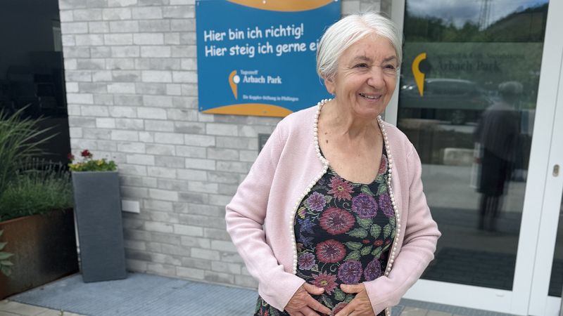 Ältere Frau steht vor einem Gebäude und lächelt. Dahinter ist ein Schild mit der Aufschrift Tagespflege zu erkennen.