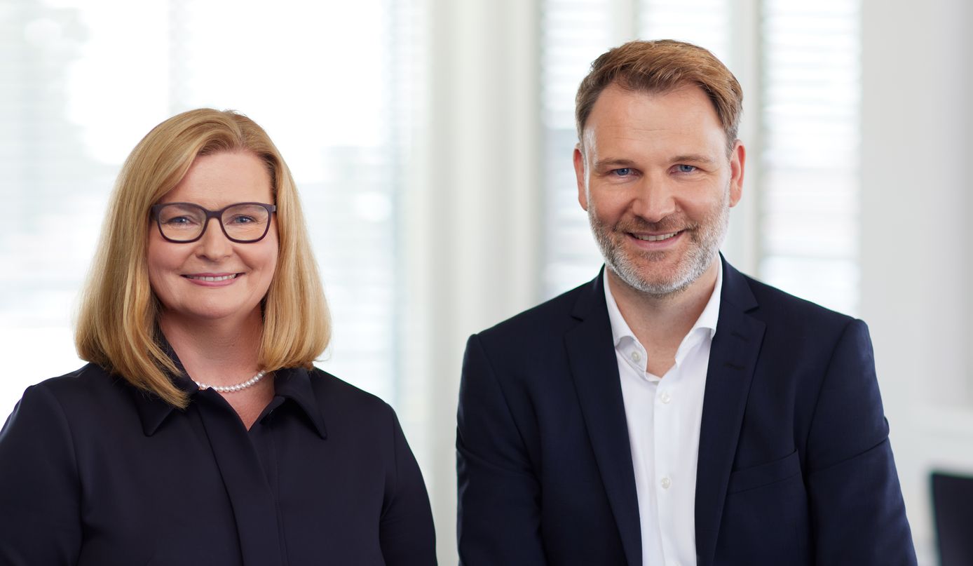 Zwei lächelnde Berufspersonen in Businesskleidung vor Bürofenstern