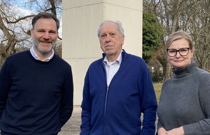 Drei Personen stehen vor einer Betonsäule, älterer Mann in der Mitte