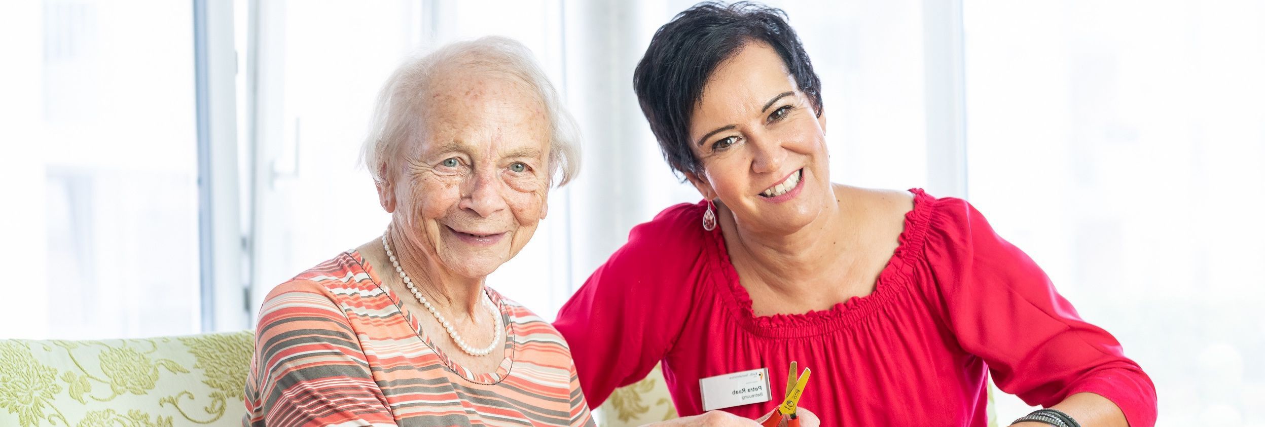 Ältere Frau und Mitarbeiterin der Stiftung lächelnd auf Sofa
