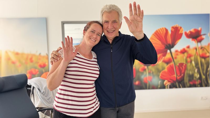 Ein älterer Mann und eine Mitarbeiterin lächeln und winken in die Kamera