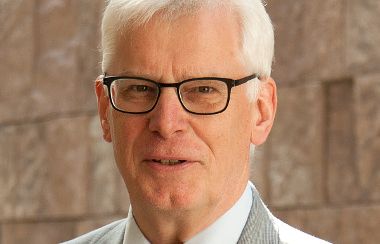 Älterer Mann mit weißem Haar, Brille, grauem Anzug und roter Krawatte