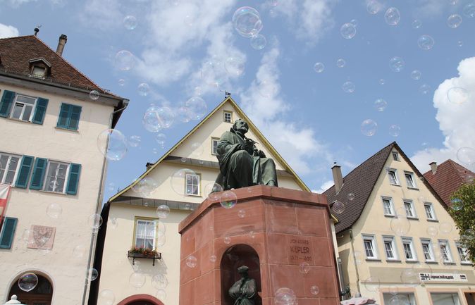 Bronzestatue auf rotem Sockel vor Häusern mit Seifenblasen