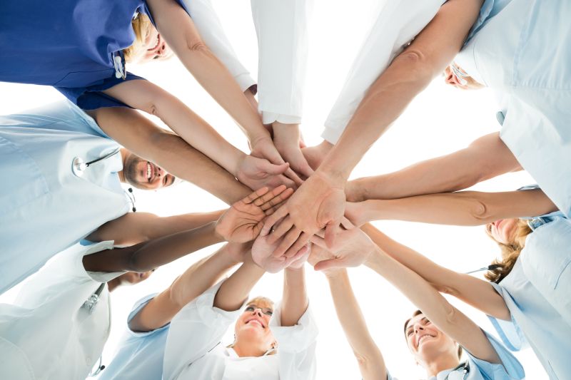 Das Bild zeigt ein motiviertes Team aus Pflegekräften und medizinischem Personal im Kreis, die ihre Hände in der Mitte aufeinanderlegen